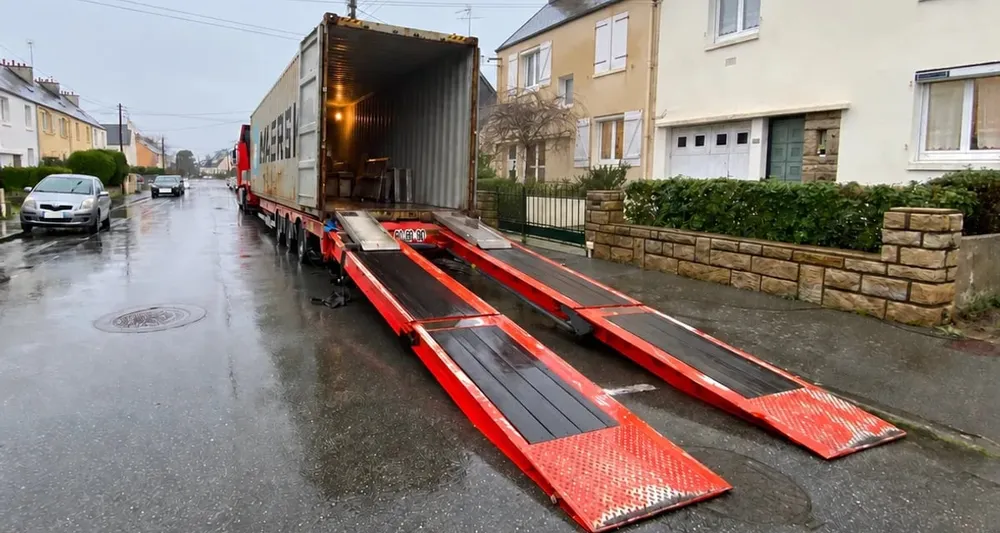 Chargement & dépôt, conteneur resté sur un châssis porte-char dans une rue résidentielle, rampes déployées jusqu’à la chaussée pour faire monter un véhicule — chantier réel IFTL
