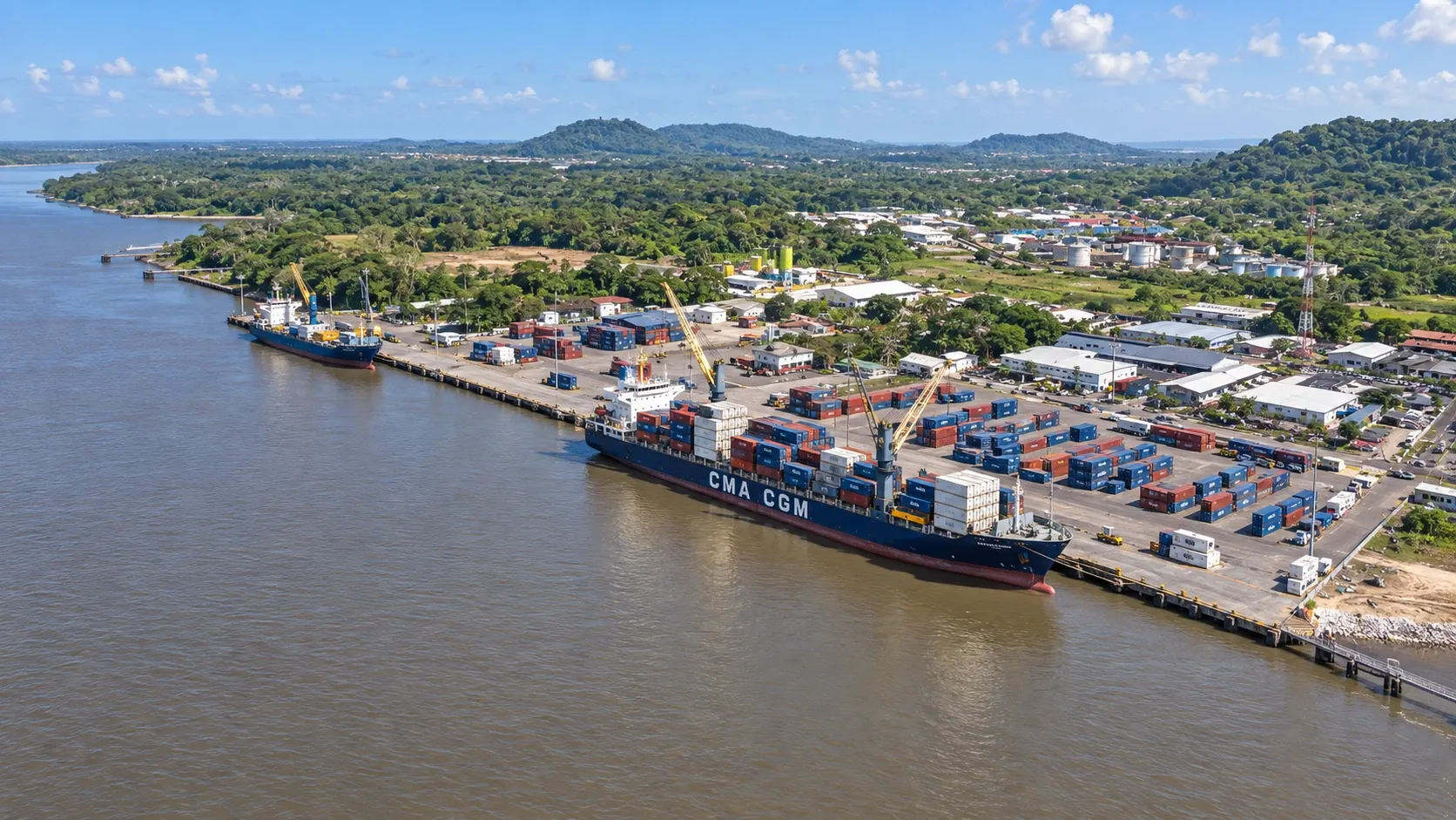 Vue aérienne d’un port fluvial : un porte-conteneurs à quai au bord d’un large fleuve, forêt tropicale à l’arrière-plan