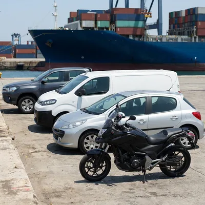Moto, voiture, fourgon et pick-up sur le quai d’un port, devant un porte-conteneurs — image d’illustration